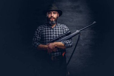 Yakışıklı sakallı avcı gezgin bir polar gömlek ve şapka tüfek bir manzara ile tutar. Karanlık bir arka plan üzerinde izole.