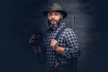 Sakallı bir avcı bir polar gömlek ve şapka portresi dürbün arkasından bir tüfekle dururken tutar. Karanlık bir arka plan üzerinde izole.