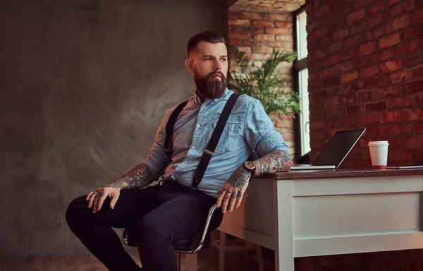 英俊的纹身时髦的衬衫和吊带坐在办公桌上的电脑 看着窗外的办公室与阁楼内部 — 图库照片