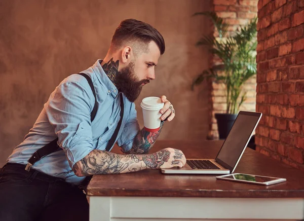 Handsome tattooed hipster in a shirt and suspenders sitting at the desk, working on a laptop, holds takeaway coffee in an office with loft interior.