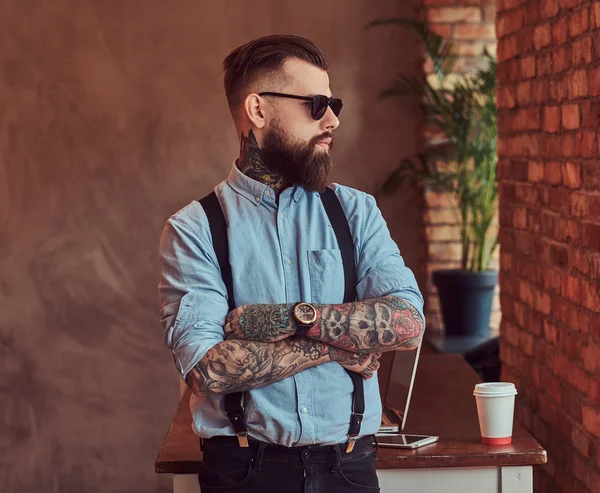 Old-fashioned tattooed hipster wearing a shirt and suspenders, in a sunglasses, standing with crossed arms near a desk with a laptop, looking out the window in an office with loft interior.