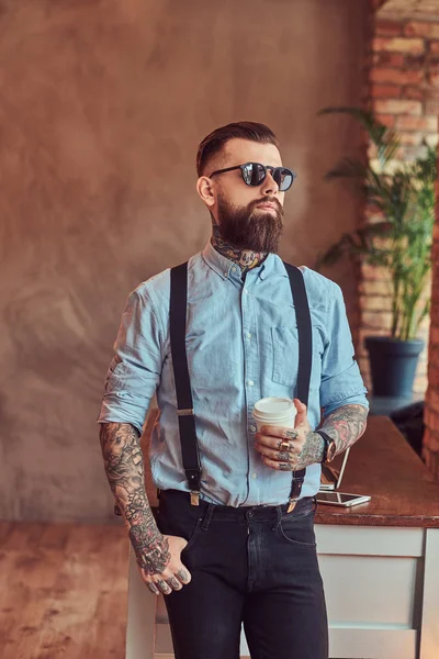 Old-fashioned tattooed hipster wearing a shirt and suspenders, in a sunglasses, standing with a cup of a coffee near a desk with a laptop, looking out the window in an office with loft interior.