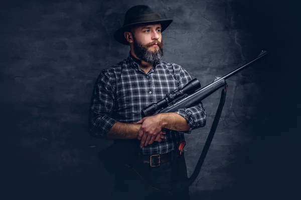 Handsome bearded hunter traveler in a fleece shirt and hat holds rifle with a sight. Isolated on a dark background.