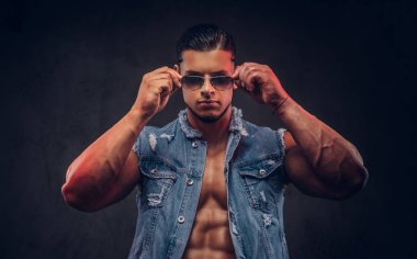 Seksi çıplak moda spor adam kot yelek ve güneş gözlüğü bir stüdyoda teşkil etmektedir. Koyu arka plan üzerinde izole.