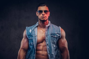 Seksi çıplak moda spor adam kot yelek ve güneş gözlüğü bir stüdyoda teşkil etmektedir. Koyu arka plan üzerinde izole.