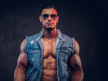 Seksi çıplak moda spor adam kot yelek ve güneş gözlüğü bir stüdyoda teşkil etmektedir. Koyu arka plan üzerinde izole.