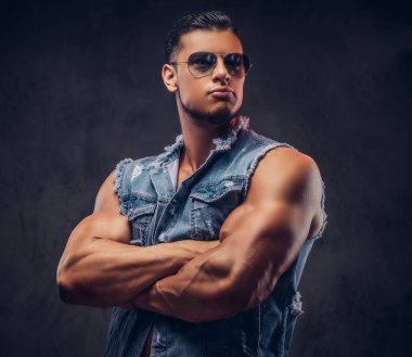 Seksi çıplak moda spor adam kot yelek ve güneş gözlüğü bir stüdyoda çapraz silah ile poz veriyor. Koyu arka plan üzerinde izole.