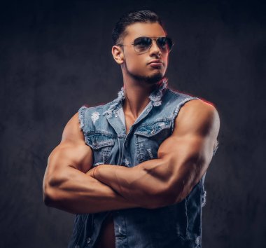 Seksi çıplak moda spor adam kot yelek ve güneş gözlüğü bir stüdyoda çapraz silah ile poz veriyor. Koyu arka plan üzerinde izole.