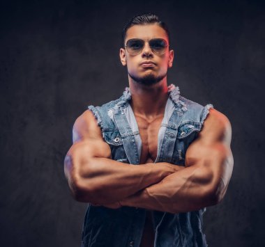 Seksi çıplak moda spor adam kot yelek ve güneş gözlüğü bir stüdyoda çapraz silah ile poz veriyor. Koyu arka plan üzerinde izole.
