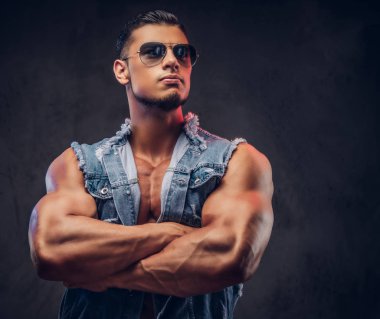 Seksi çıplak moda spor adam kot yelek ve güneş gözlüğü bir stüdyoda çapraz silah ile poz veriyor. Koyu arka plan üzerinde izole.