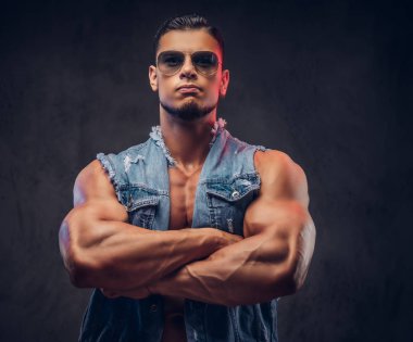 Seksi çıplak moda kendine güvenen erkek denim yelek ve güneş gözlüğü kaslı vücudu olan bir stüdyoda çapraz silah ile poz veriyor. Koyu arka plan üzerinde izole.