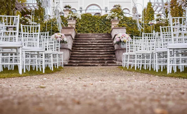 Place for wedding ceremony with wedding arch decorated with flowers and white chairs on each side of archway outdoors. Preparation for wedding ceremony outdoors near lake.