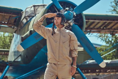 Tam bir uçuş yiğit yakışıklı pilot ayakta retro askeri uçak yakınındaki Dişli ve uzak görünüyor.