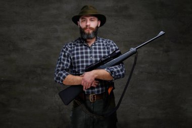 Yakışıklı sakallı avcı gezgin bir polar gömlek ve şapka tüfek bir manzara ile tutar. Karanlık bir arka plan üzerinde izole.