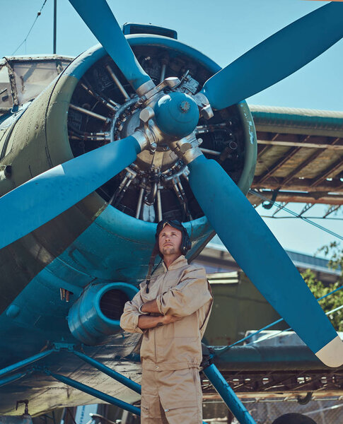 Handsome pilot in a full flight gear standing with crossed arms near military airplane.