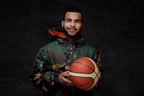 Happy young African-American street basketball player in a camouflage jacket. Isolated over dark background. 