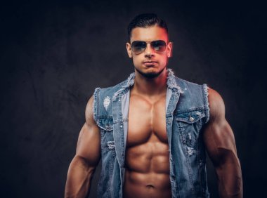 Seksi çıplak moda spor adam kot yelek ve güneş gözlüğü bir stüdyoda teşkil etmektedir. Koyu arka plan üzerinde izole.