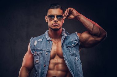 Seksi çıplak moda kendine güvenen erkek denim yelek ve güneş gözlüğü kaslı vücudu olan bir stüdyoda teşkil etmektedir. Koyu arka plan üzerinde izole.