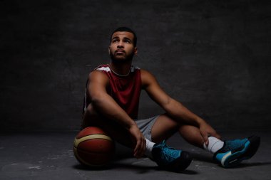 Spor giyim topuyla bir katta oturan genç Afro-Amerikan basketbol oyuncusu. Koyu arka plan üzerinde izole. 