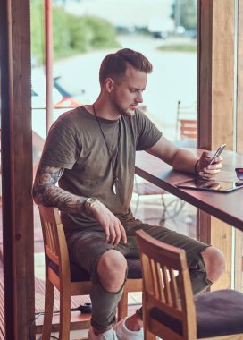 Bir smartphone cep telefonu ile mesaj okuma yakışıklı şık hipster yol kenarına yerleştirilen bir kafede bir masada oturur.