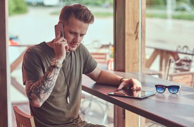 Konuşmak smartphone yakışıklı şık hipster yol kenarına yerleştirilen bir kafede bir masada oturur.