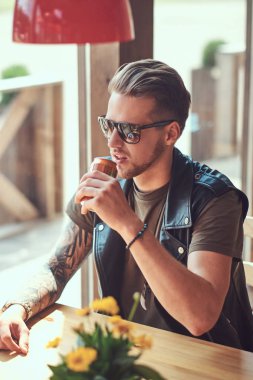 Yenilikçi bir şık saç ve sakal ile yol kenarına yerleştirilen bir kafede bir masada oturuyor, sıcak bir günde bir soda içer.