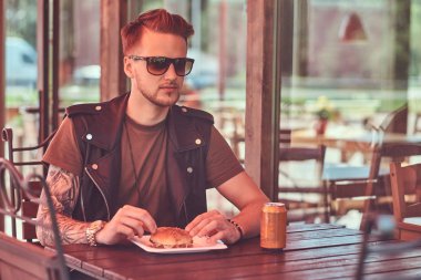 Yakışıklı şık hipster adam portresi bir hamburger ve soda sipariş bir masada oturuyor, yol kenarına yerleştirilen bir café'de akşam