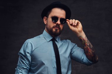 Yakışıklı, dövmeli bir orta yaşlı adam sakal ve saç modeli ile portre resmi elbise ve şapka giymiş, güneş gözlüğü üzerinde el ile poz. Bir stüdyoda dokulu koyu arka plan üzerinde izole.