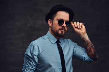 Yakışıklı, dövmeli bir orta yaşlı adam sakal ve saç modeli ile portre resmi elbise ve şapka giymiş, güneş gözlüğü üzerinde el ile poz. Bir stüdyoda dokulu koyu arka plan üzerinde izole.