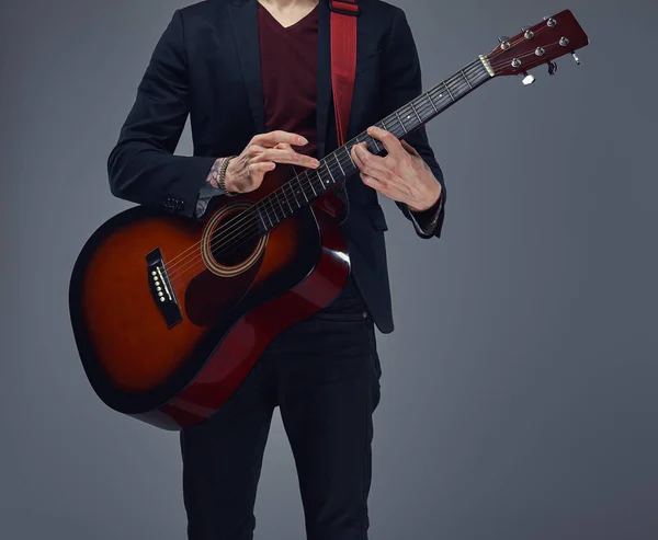 Handsome young musician with stylish hair in elegant clothes, playing on an acoustic guitar. Isolated on a gray background.