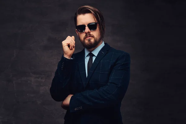 Pensive handsome fashionable middle-aged man with beard and hairstyle dressed in an elegant formal suit and sunglasses on a textured dark background in studio.