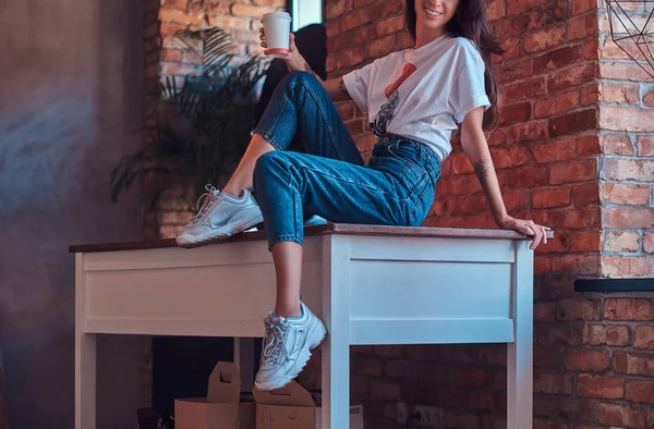 一个凉爽的现代女孩的形象 穿着白色的顶部和牛仔裤坐在桌子上 一杯外卖咖啡与阁楼内部的工作室 — 图库照片