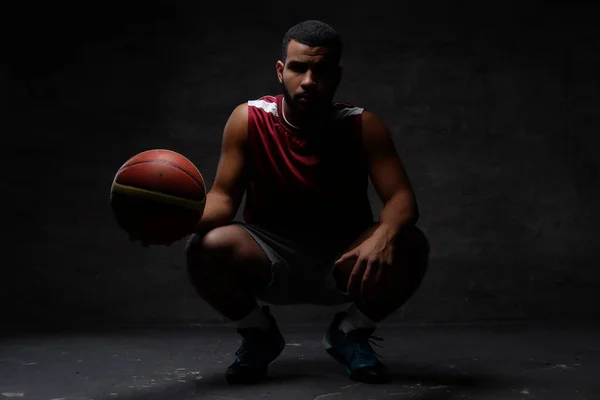 スポーツ ウエア ボールとのしゃがむでアフリカ系アメリカ人のバスケット ボール選手 暗い背景上分離 — ストック写真