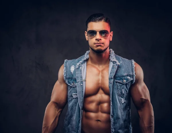 Sexy naked fashionable athletic man in a denim vest and sunglasses poses in a studio. — Stock Photo, Image