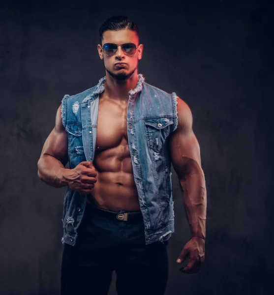 Sexy homme athlétique nu à la mode dans un gilet en denim et des lunettes de soleil pose dans un studio . — Photo