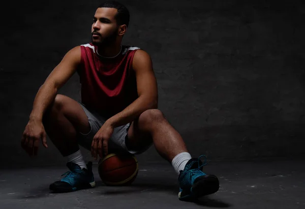 Young African-American basketball player in sportswear sitting on a ball on a dark background. — Stock Photo, Image