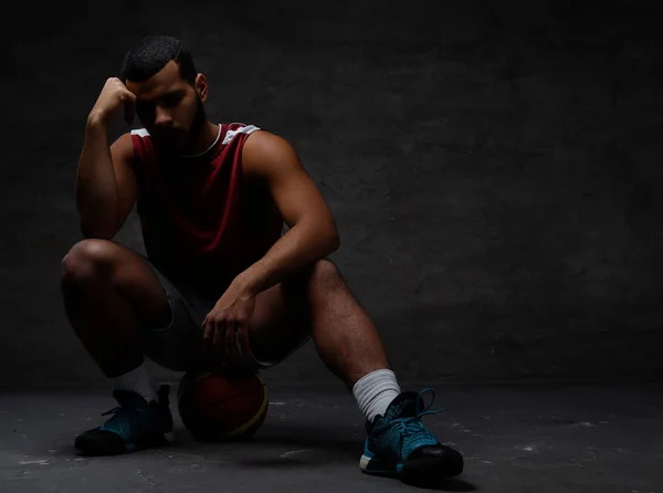 Jeune joueur de basket afro-américain pensif en tenue de sport assis sur une balle sur fond sombre . — Photo