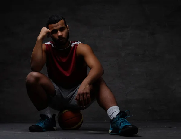 Nachdenklicher junger afrikanisch-amerikanischer Basketballspieler in Sportbekleidung auf einem Ball vor dunklem Hintergrund sitzend. — Stockfoto