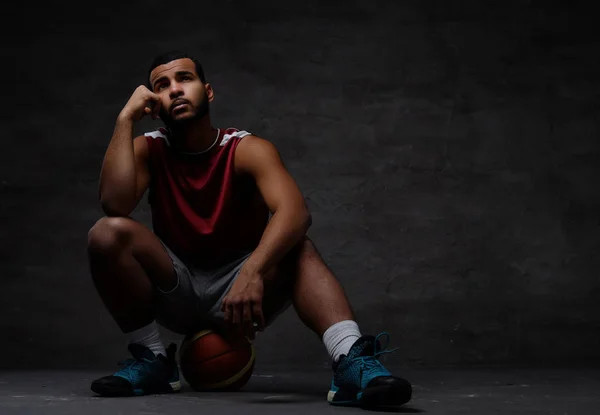 Nachdenklicher junger afrikanisch-amerikanischer Basketballspieler in Sportbekleidung auf einem Ball vor dunklem Hintergrund sitzend. — Stockfoto