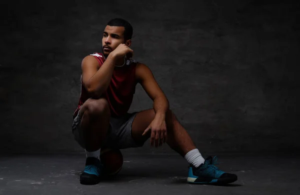 Pensive young African-American basketball player in sportswear sitting on a ball over dark background. — Stock Photo, Image