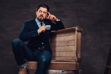 Dalgın orta yaş işadamı şık saç ve sakal ile kot pantolon, ceket ve kravat, ahşap kutular üzerinde oturan giymiş ve bir fincan kahve bir stüdyoda tutar.