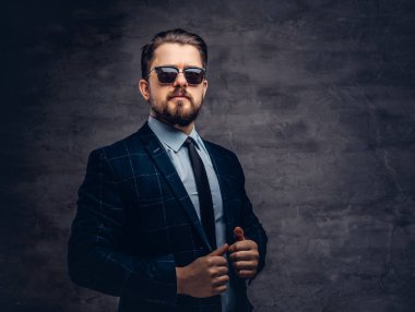 Giyinmiş emin yakışıklı şık orta yaşlı adam sakal ve saç modeli ile zarif bir resmi takım elbise ve Studio karanlık bir arka plan üzerinde güneş gözlüğü.