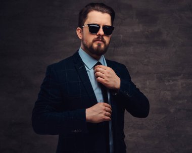 Giyinmiş emin yakışıklı şık orta yaşlı adam sakal ve saç modeli ile zarif bir resmi takım elbise ve Studio karanlık bir arka plan üzerinde güneş gözlüğü.
