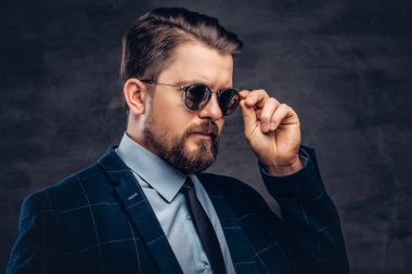 Giyinmiş emin yakışıklı şık orta yaşlı adam sakal ve saç modeli ile zarif bir resmi takım elbise ve Studio karanlık bir arka plan üzerinde güneş gözlüğü.