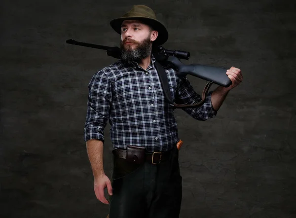 Bearded hunter in a fleece shirt and hat holds on his shoulder a rifle with sight, looking away.