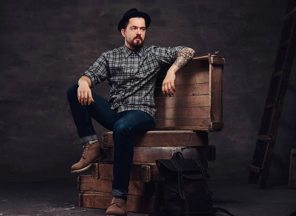 Full body portrait of a tattoed middle age hipster man dressed in a checkered shirt, jeans and hat, sitting on wooden boxes in a studio.