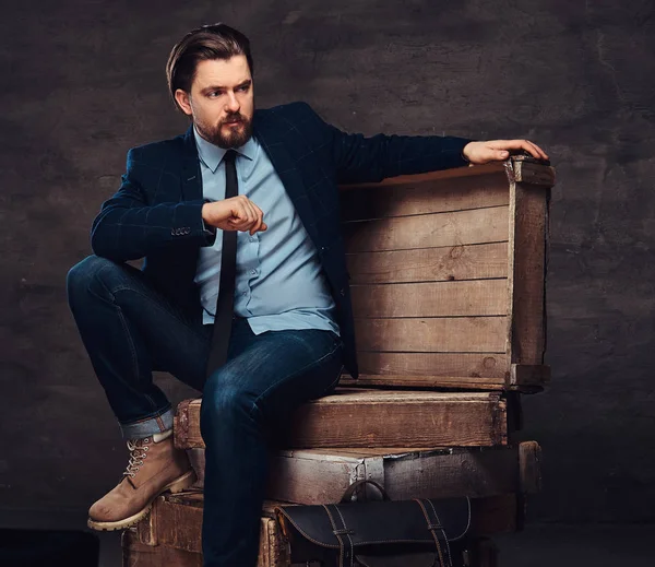 Portrét podnikatel středního věku s elegantní vlasy a vousy na sobě džíny, sako a kravatu, sedí na dřevěných beden ve studiu. — Stock fotografie