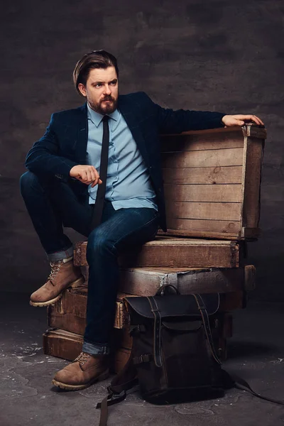 Portrét podnikatel středního věku s elegantní vlasy a vousy na sobě džíny, sako a kravatu, sedí na dřevěných beden ve studiu. — Stock fotografie