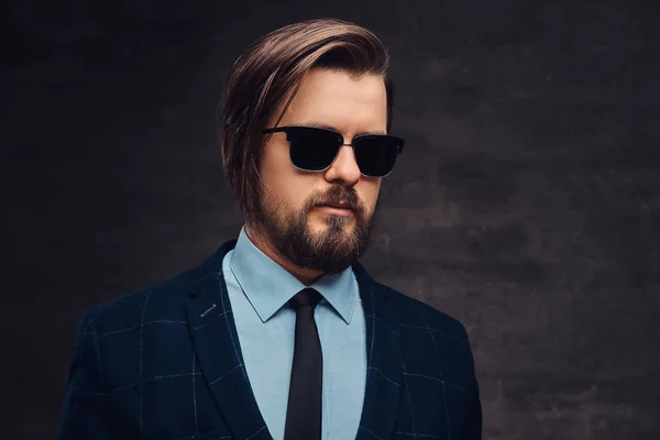 Close-up portrait of a handsome fashionable middle-aged man with beard and hairstyle dressed in an elegant formal suit and sunglasses. Isolated on a textured dark background in studio. — Stock Photo, Image