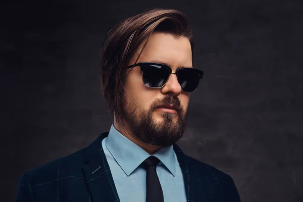 Gros plan portrait d'un bel homme à la mode d'âge moyen avec barbe et coiffure vêtu d'un élégant costume formel et de lunettes de soleil. Isolé sur un fond sombre texturé en studio . — Photo